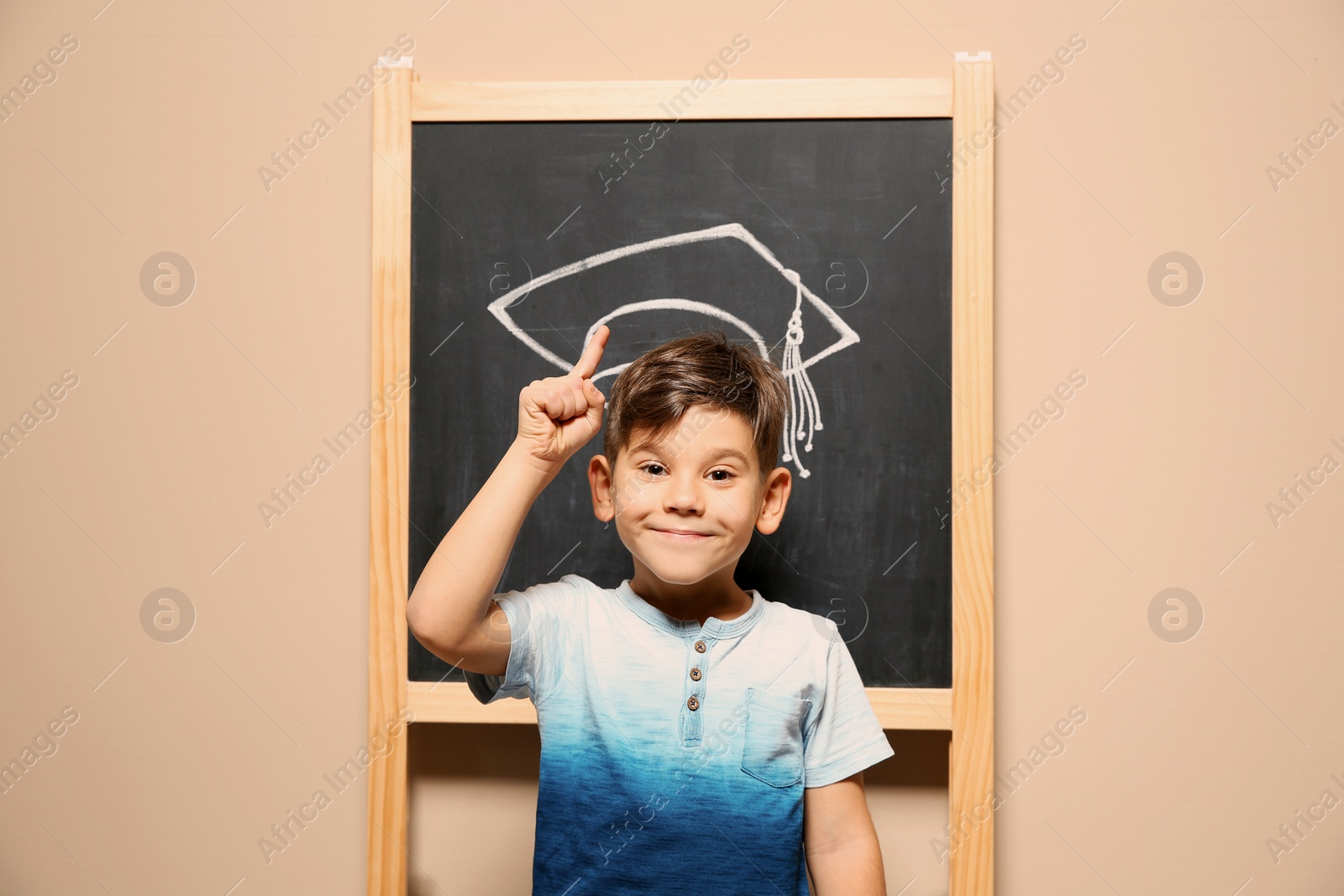 Photo of Cute little child standing at blackboard with chalk drawn academic cap. Education concept