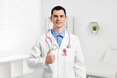 Photo of Portrait of smiling mammologist with stethoscope and pink ribbon showing thumb up in hospital. Breast cancer awareness