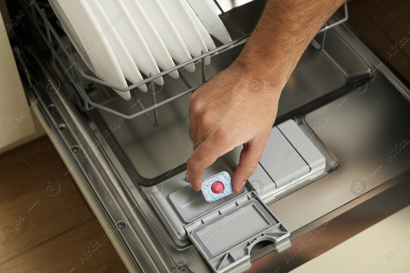 Photo of Man putting detergent tablet into open dishwasher indoors, closeup