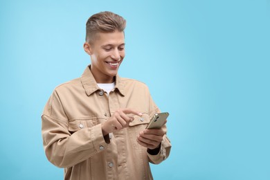 Photo of Happy young man sending message via smartphone on light blue background. Space for text