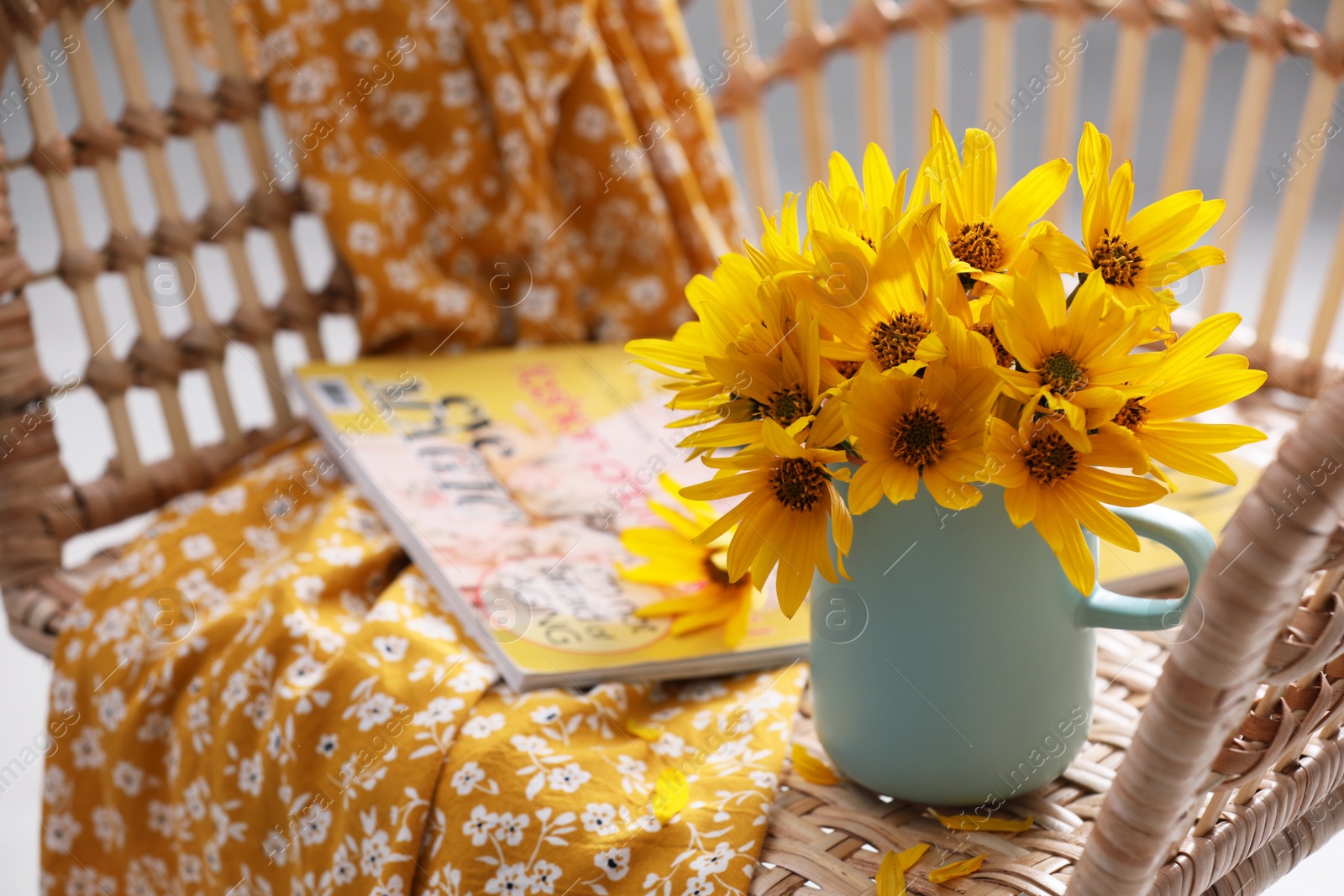 Photo of Beautiful bright flowers in light blue cup near magazine and fabric on rattan armchair. Space for text