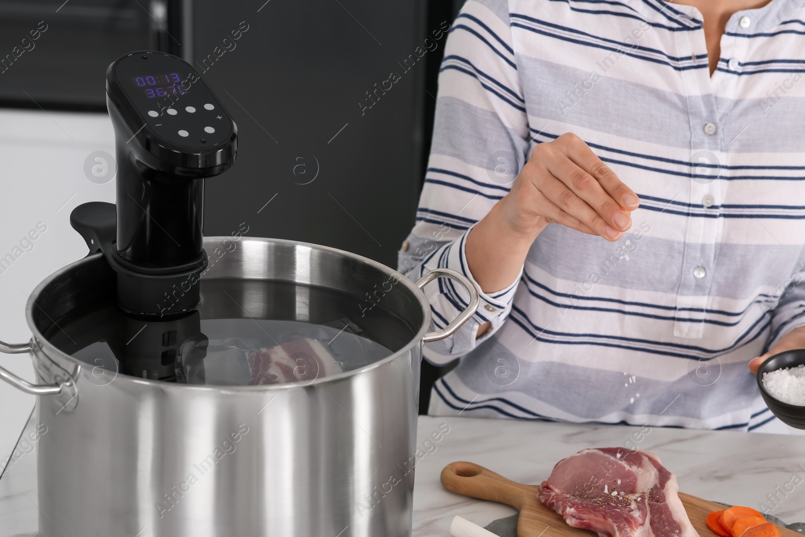 Photo of Woman salting meat near pot with sous vide cooker at table, closeup. Thermal immersion circulator