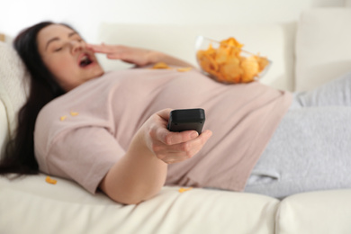 Photo of Lazy overweight woman with chips watching TV at home, focus on remote control