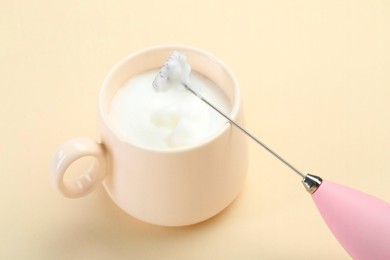 Photo of Mini mixer (milk frother) and cup of whipped milk on beige background, closeup