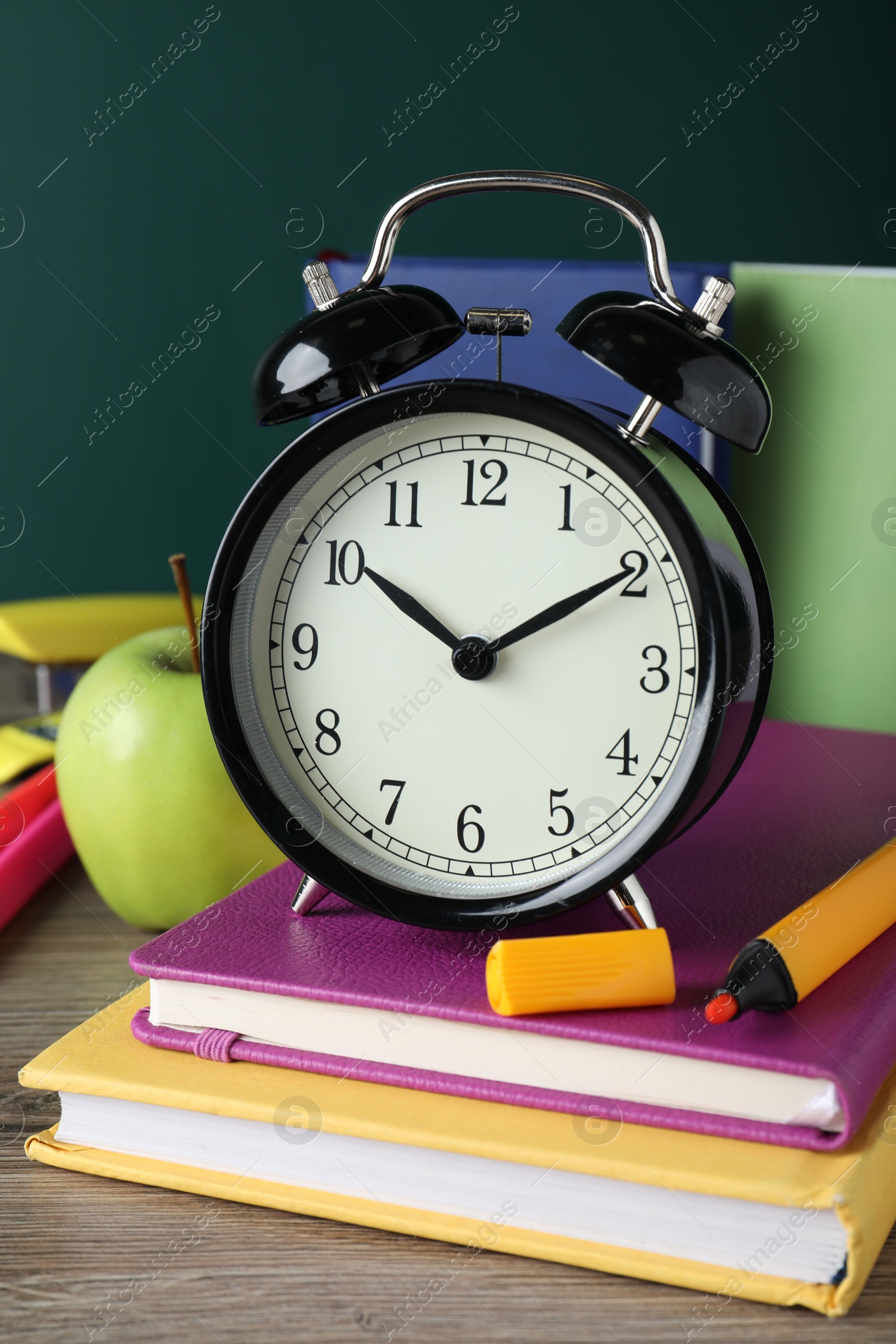 Photo of Alarm clock and different stationery on wooden table near green chalkboard. School time