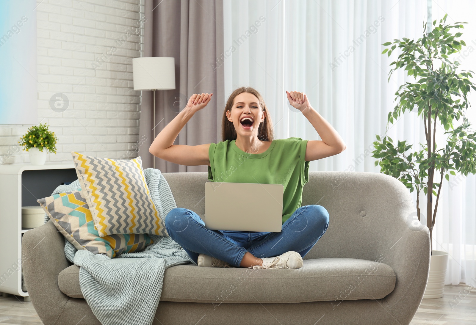 Photo of Emotional young woman with laptop celebrating victory on sofa at home