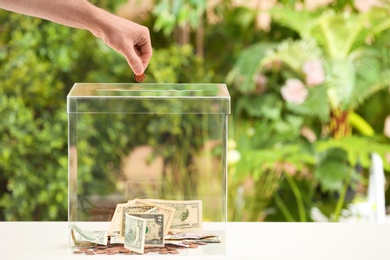 Man putting coins into donation box on table against blurred background, closeup. Space for text