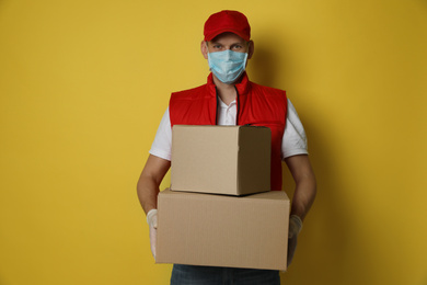 Photo of Courier in protective mask and gloves holding cardboard boxes on yellow background. Delivery service during coronavirus quarantine