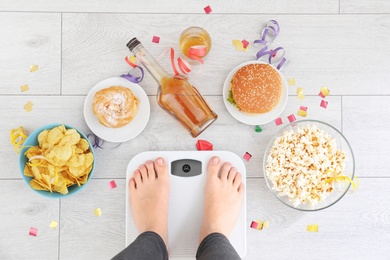 Photo of Woman standing on scales surrounded by different food and alcohol after party indoors, top view