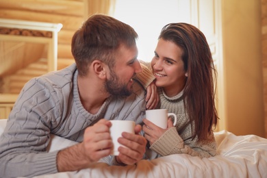 Young couple in warm sweaters with cups of hot drink on bed at home