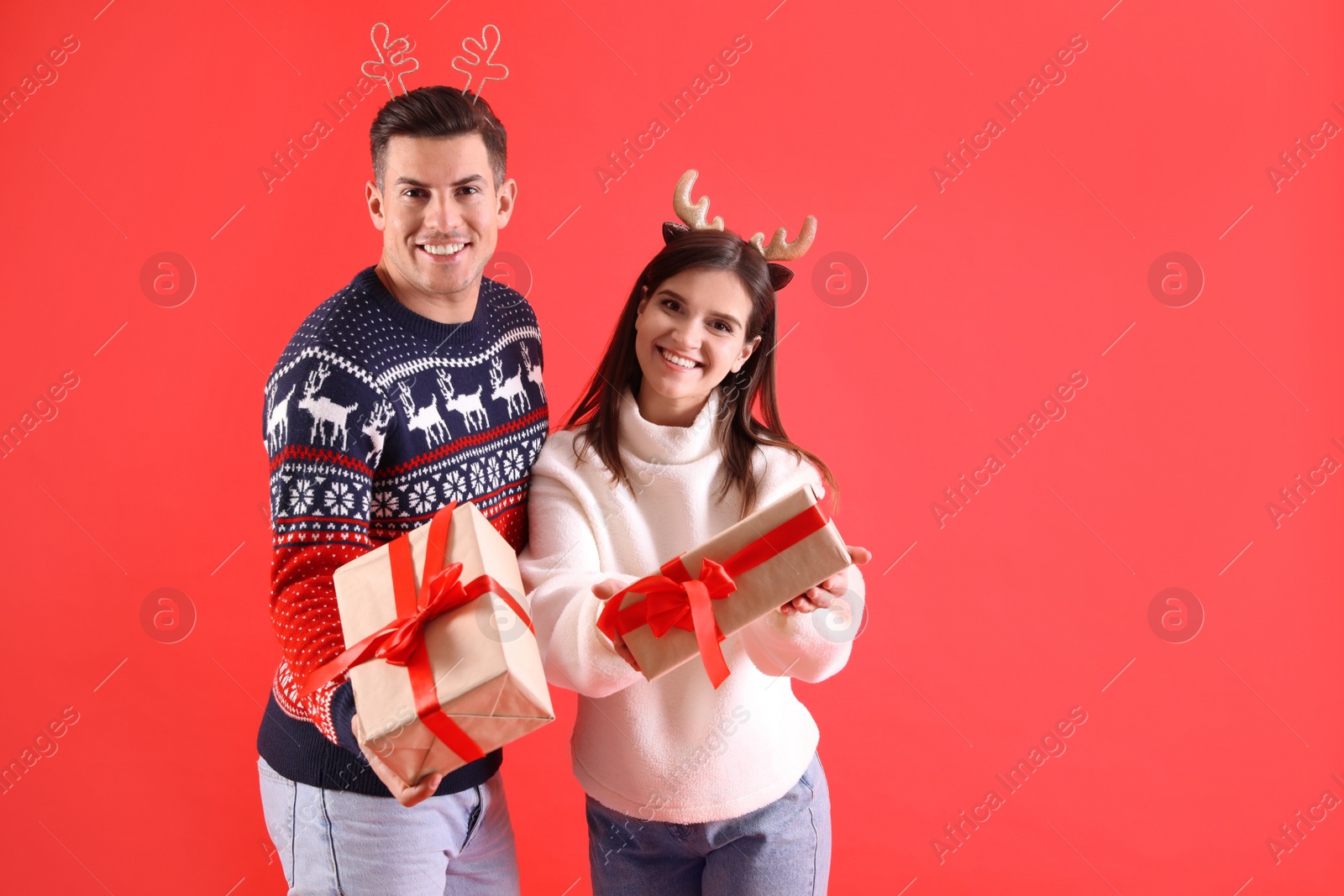 Photo of Beautiful happy couple in Christmas headbands and sweaters holding gifts on red background