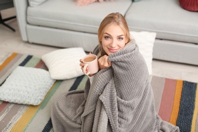 Beautiful young woman wrapped in plaid sitting with cup of coffee on floor at home. Winter atmosphere