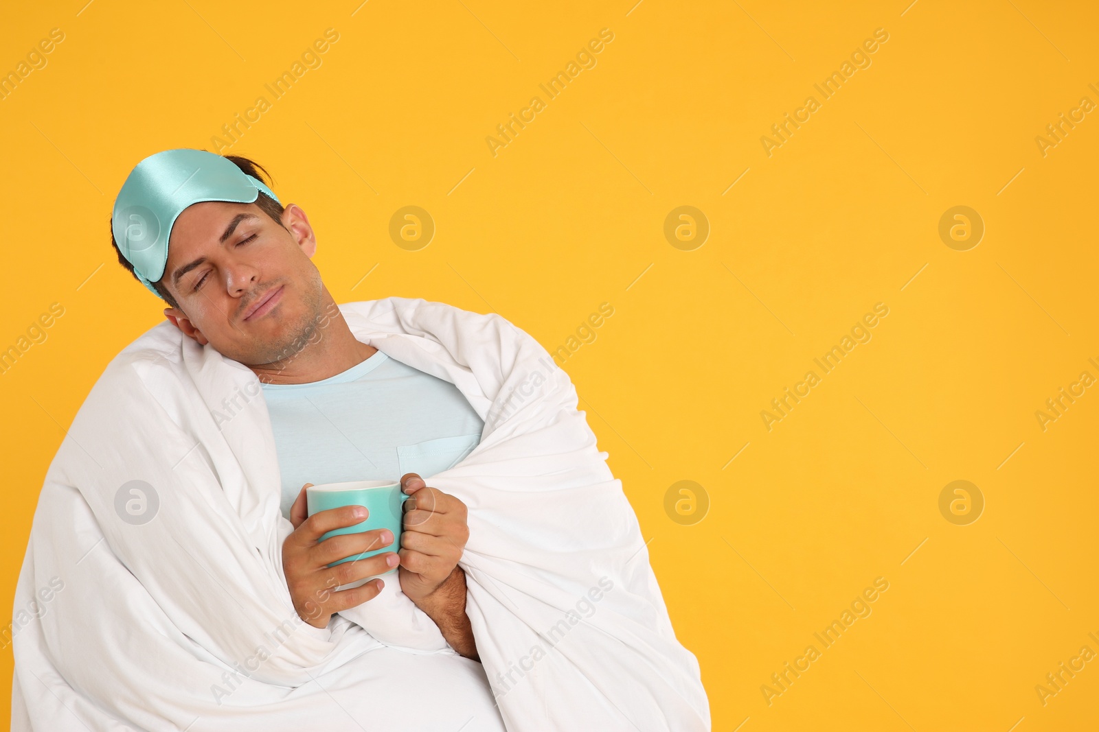 Photo of Man in sleeping mask wrapped with blanket holding cup on yellow background. Space for text