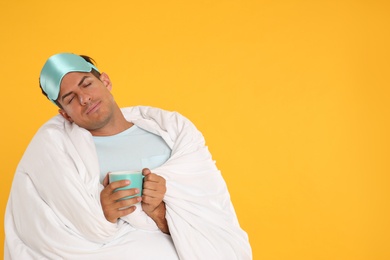 Photo of Man in sleeping mask wrapped with blanket holding cup on yellow background. Space for text