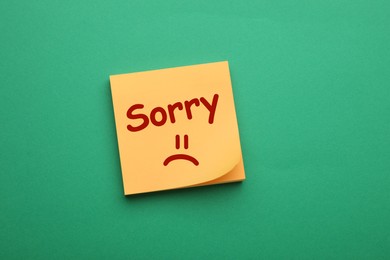 Image of Apology. Sticky note with word Sorry and drawn sad face on green background, top view