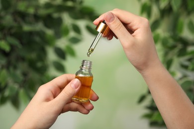 Woman dripping CBD oil from pipette into bottle against blurred green background, closeup