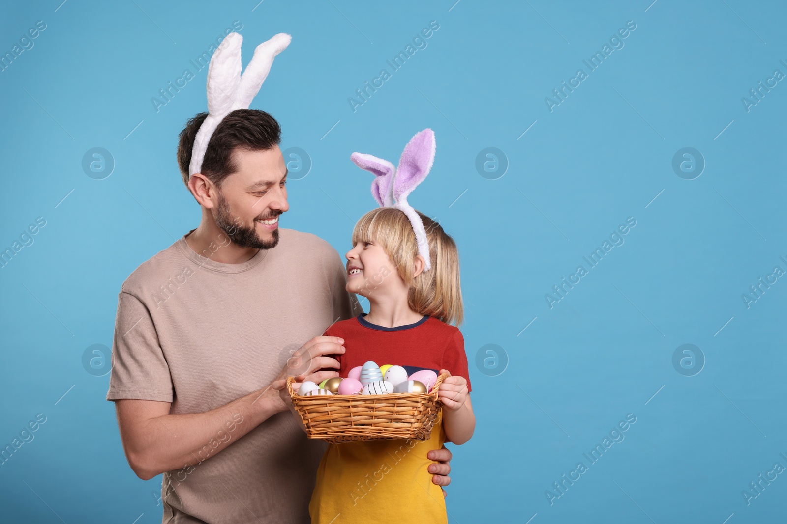 Photo of Father and son in bunny ears headbands with wicker basket of painted Easter eggs on turquoise background. Space for text