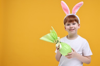 Photo of Easter celebration. Cute little boy with bunny ears and wrapped egg on orange background. Space for text