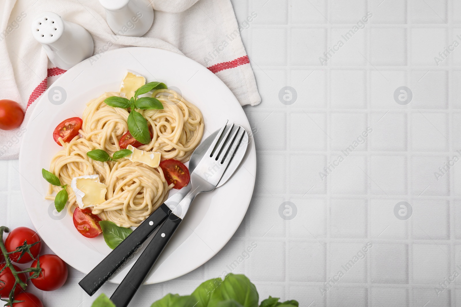 Photo of Delicious pasta with brie cheese, tomatoes, basil and cutlery on white tiled table, flat lay. Space for text