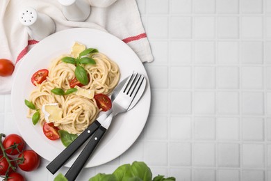 Photo of Delicious pasta with brie cheese, tomatoes, basil and cutlery on white tiled table, flat lay. Space for text