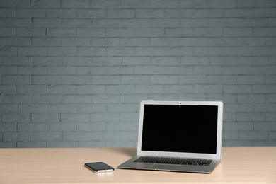 Photo of Modern laptop with blank screen and mobile phone on table against brick wall