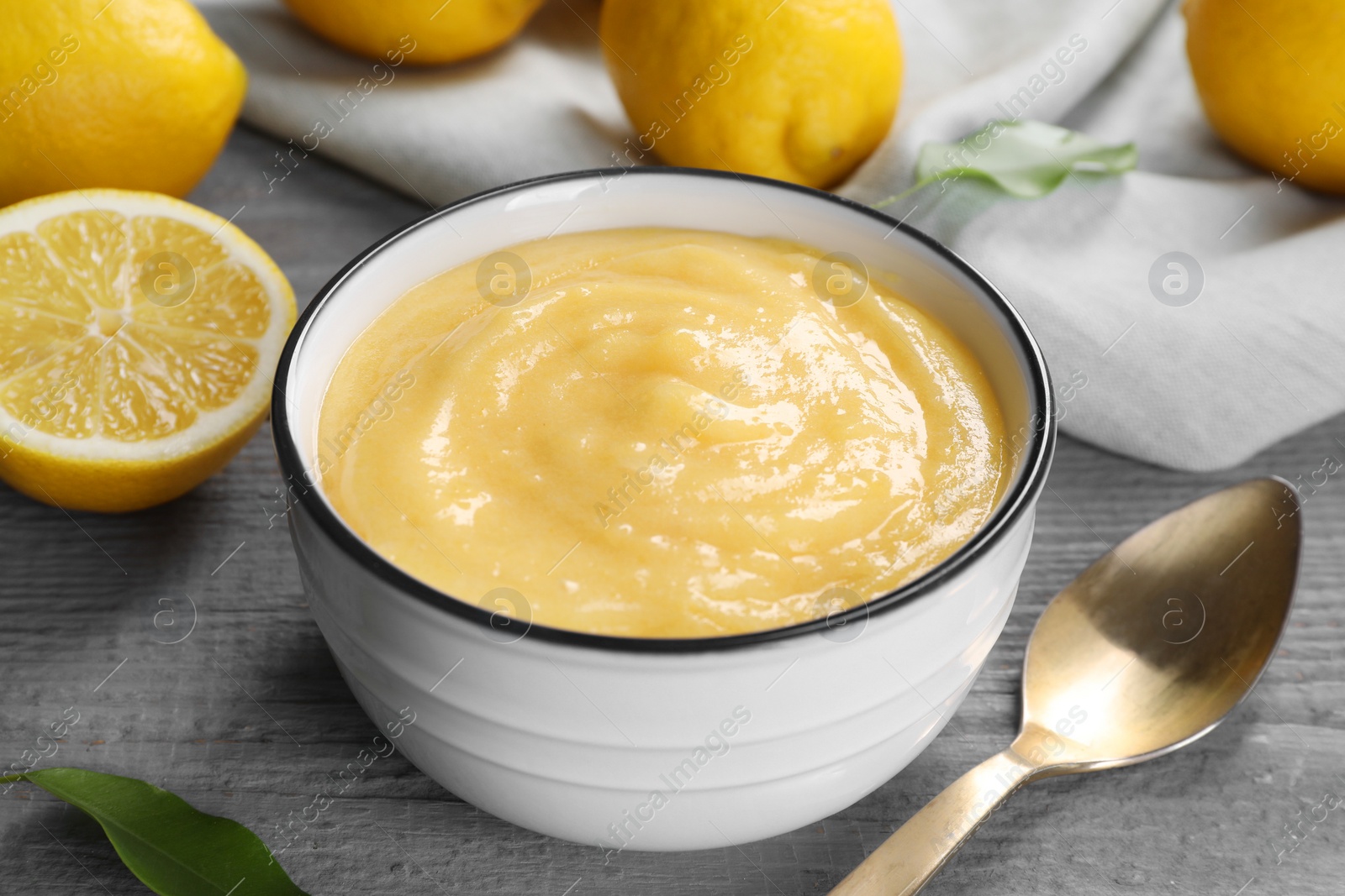 Photo of Delicious lemon curd in bowl, fresh citrus fruits and spoon on grey wooden table, closeup