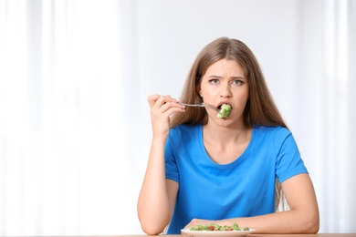 Portrait of unhappy woman eating broccoli salad at table on light background