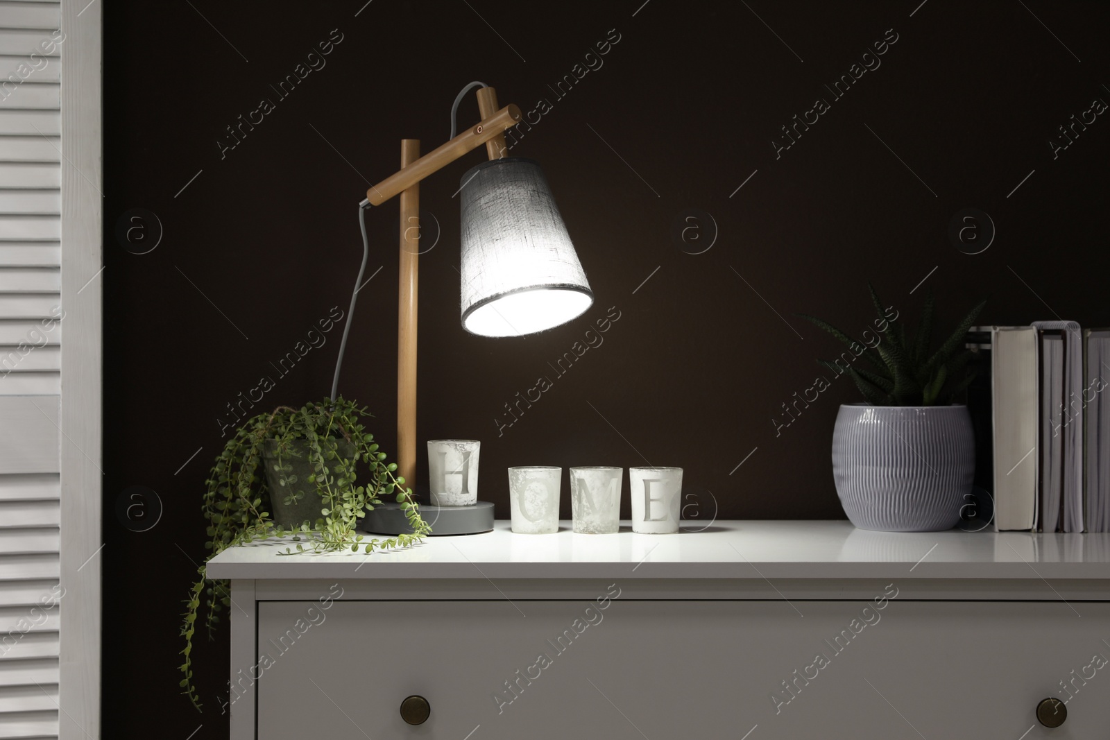 Photo of Stylish lamp, books and green plants on white chest of drawers near brown wall indoors. Interior design