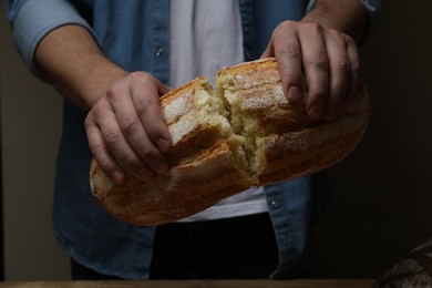 Photo of Man breaking loaf of fresh bread on dark background, closeup