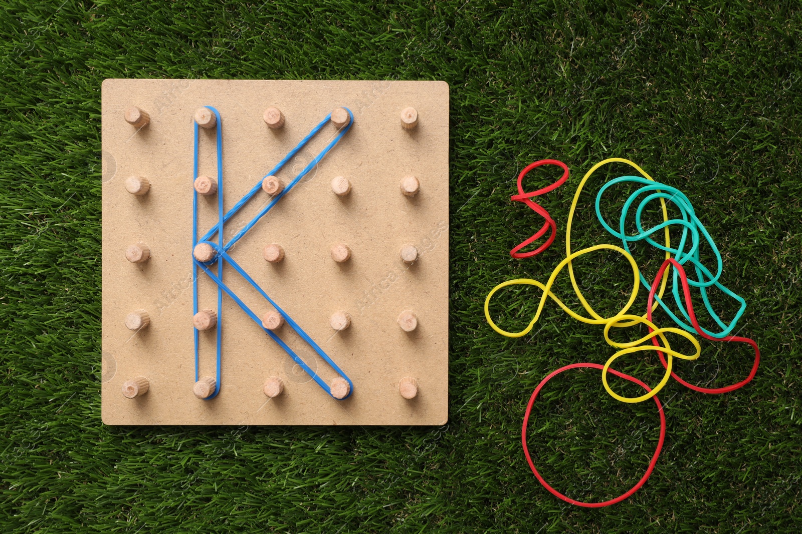 Photo of Wooden geoboard with letter K made of rubber bands on artificial grass, flat lay. Educational toy for motor skills development