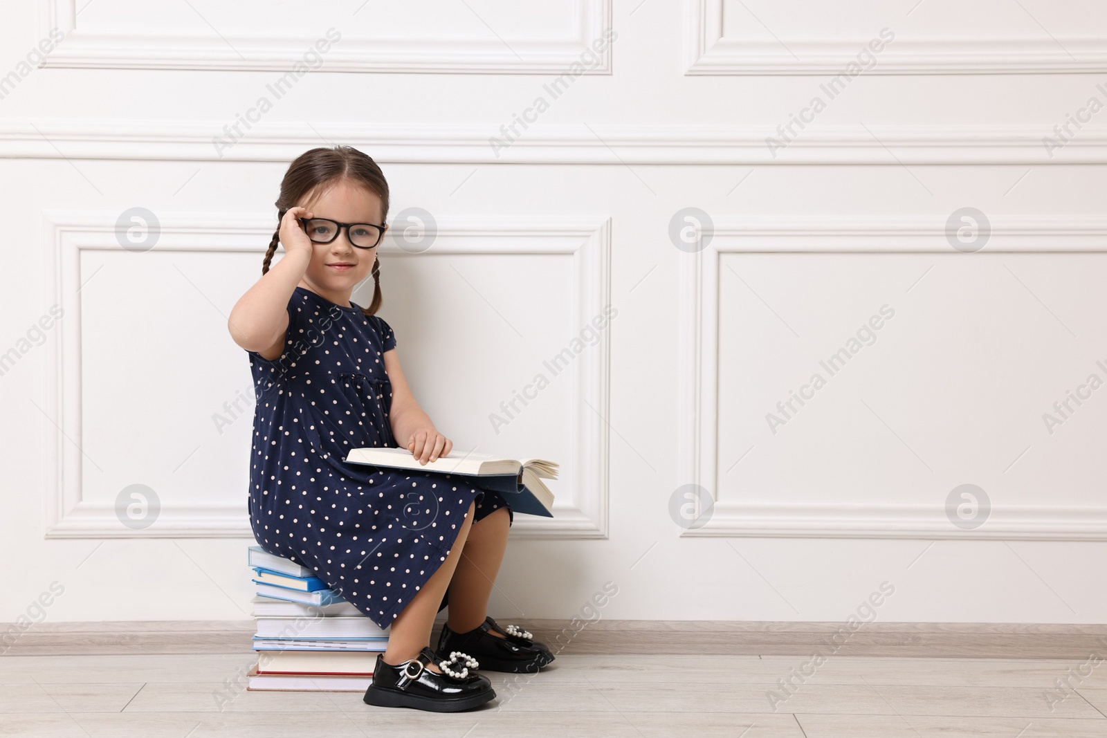 Photo of Cute little girl in glasses sitting on stack of books near white wall. Space for text