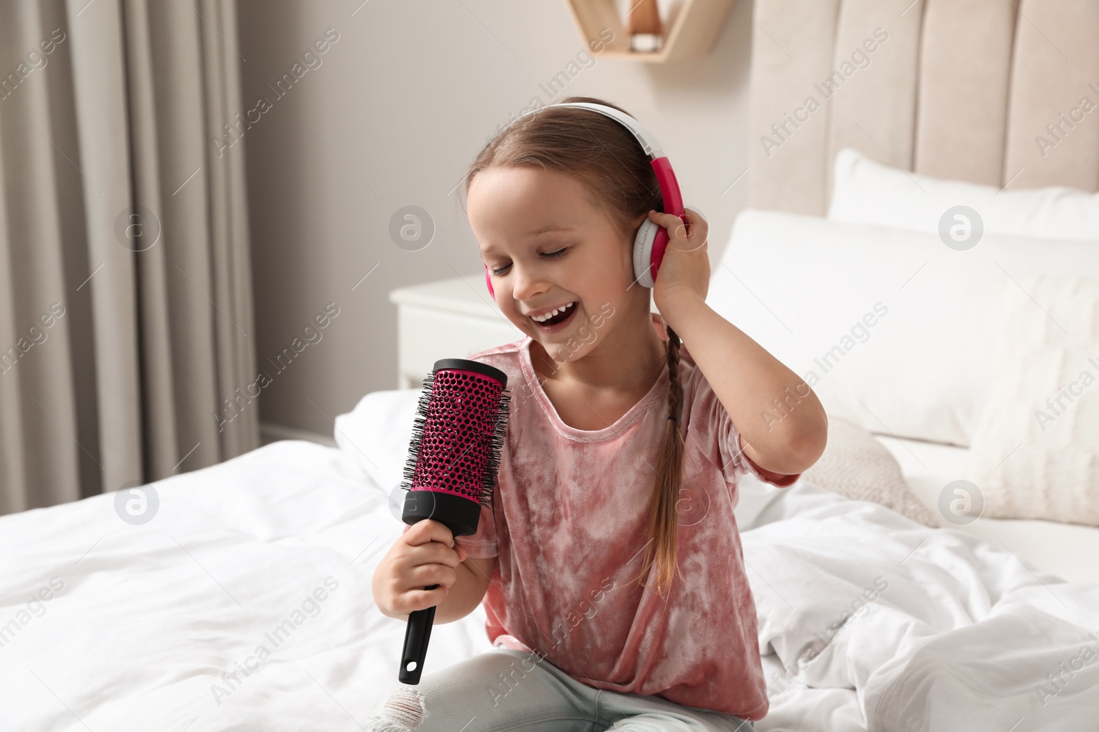 Photo of Cute little girl in headphones with hairbrush singing on bed at home