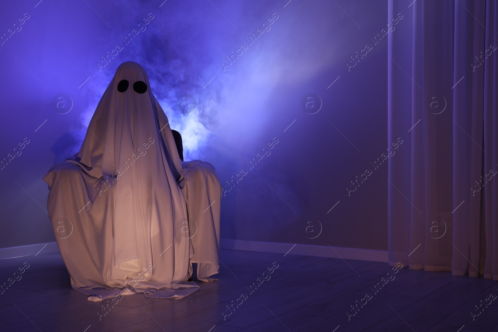 Photo of Creepy ghost. Woman covered with sheet sitting in armchair in blue light, space for text