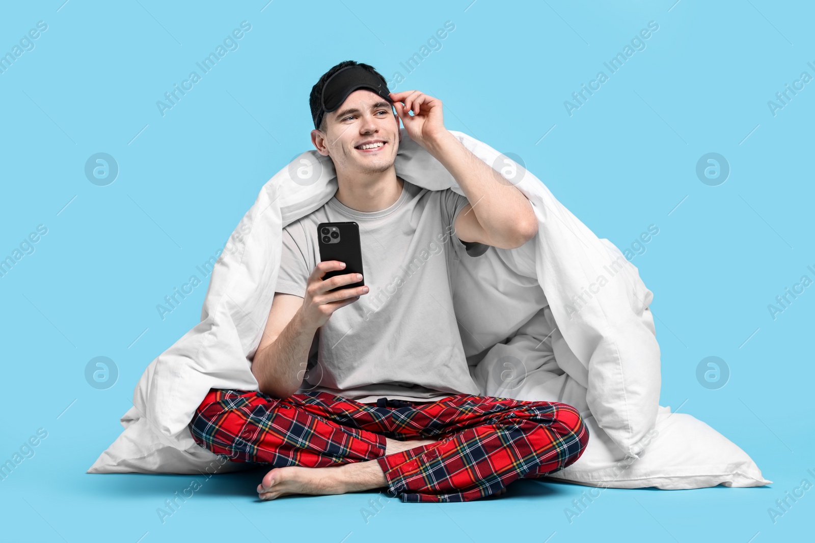 Photo of Happy man in pyjama wrapped in blanket using smartphone on light blue background