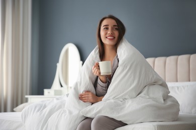 Photo of Beautiful young woman wrapped with soft blanket holding cup of hot drink on bed at home