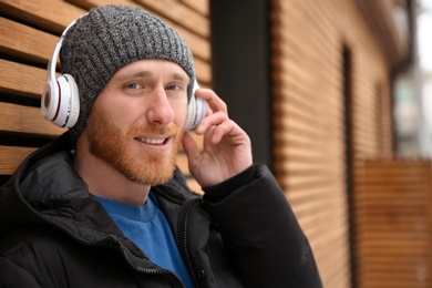 Young man listening to music with headphones near wooden wall. Space for text