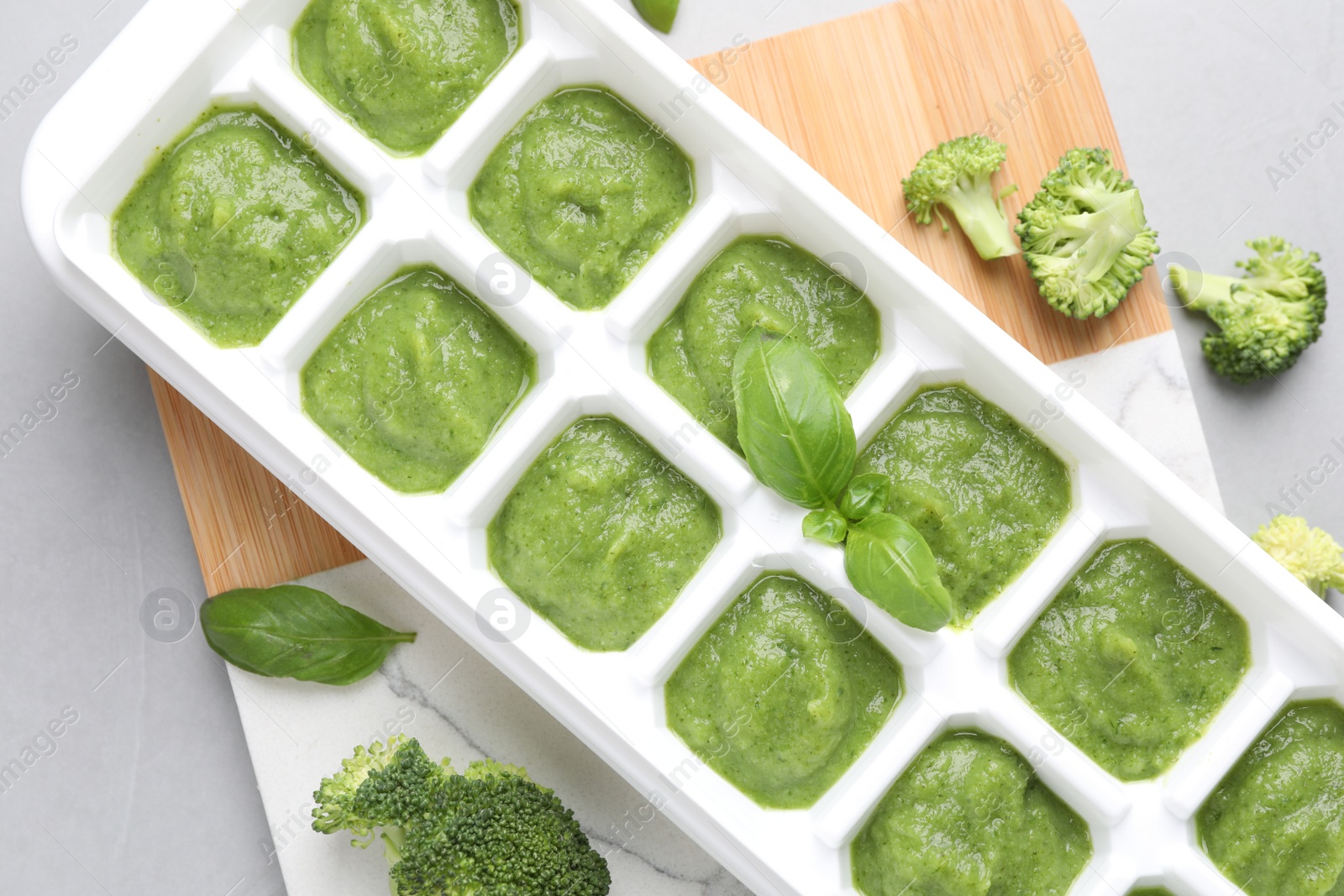 Photo of Broccoli puree in ice cube tray and ingredients on grey table, flat lay. Ready for freezing