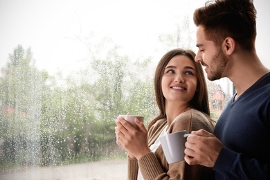 Happy young couple with cups near window indoors on rainy day