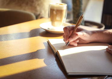 Woman with notebook and cup of latte at wooden table, closeup