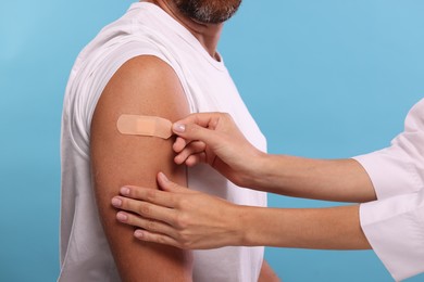 Nurse sticking plaster on man's arm after vaccination against light blue background, closeup