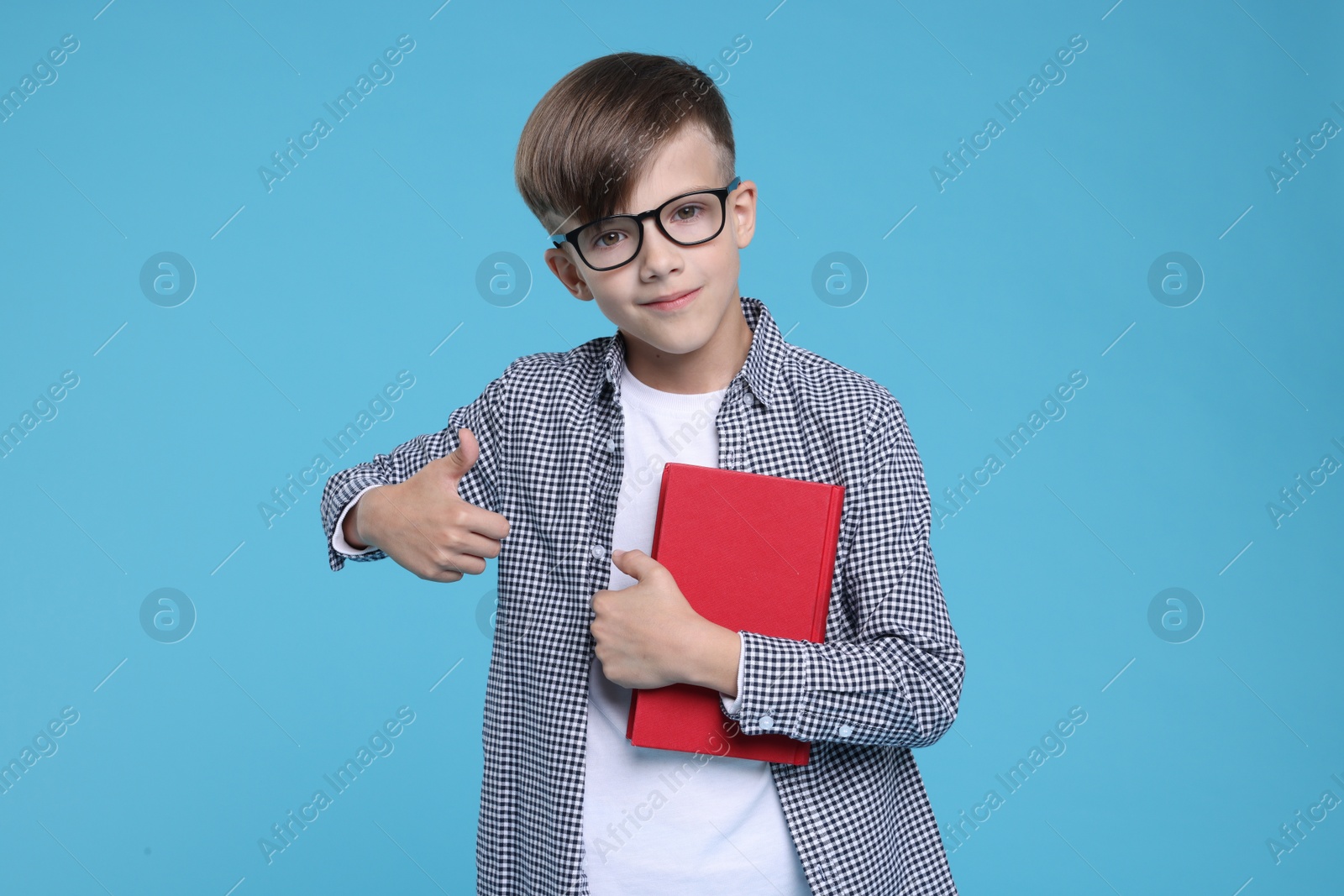 Photo of Cute schoolboy in glasses with book showing thumbs up on light blue background