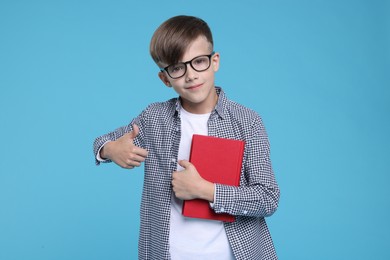 Cute schoolboy in glasses with book showing thumbs up on light blue background