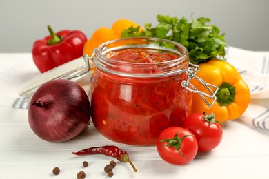 Glass jar of delicious canned lecho and fresh ingredients on white wooden table, closeup