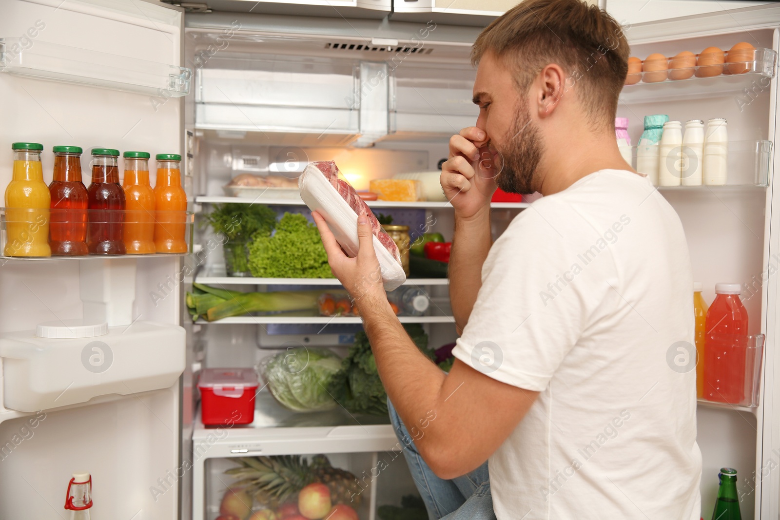 Photo of Young man with bad smelling meat near refrigerator in kitchen