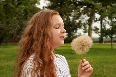 Cute girl with beautiful red hair blowing large dandelion in park. Allergy free concept