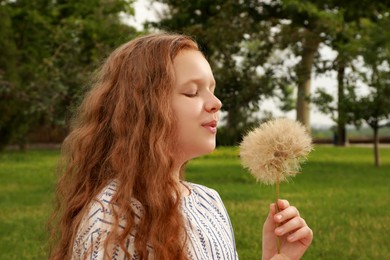 Photo of Cute girl with beautiful red hair blowing large dandelion in park. Allergy free concept