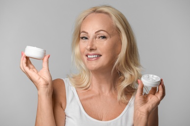 Portrait of beautiful mature woman with perfect skin holding jars of cream on grey background