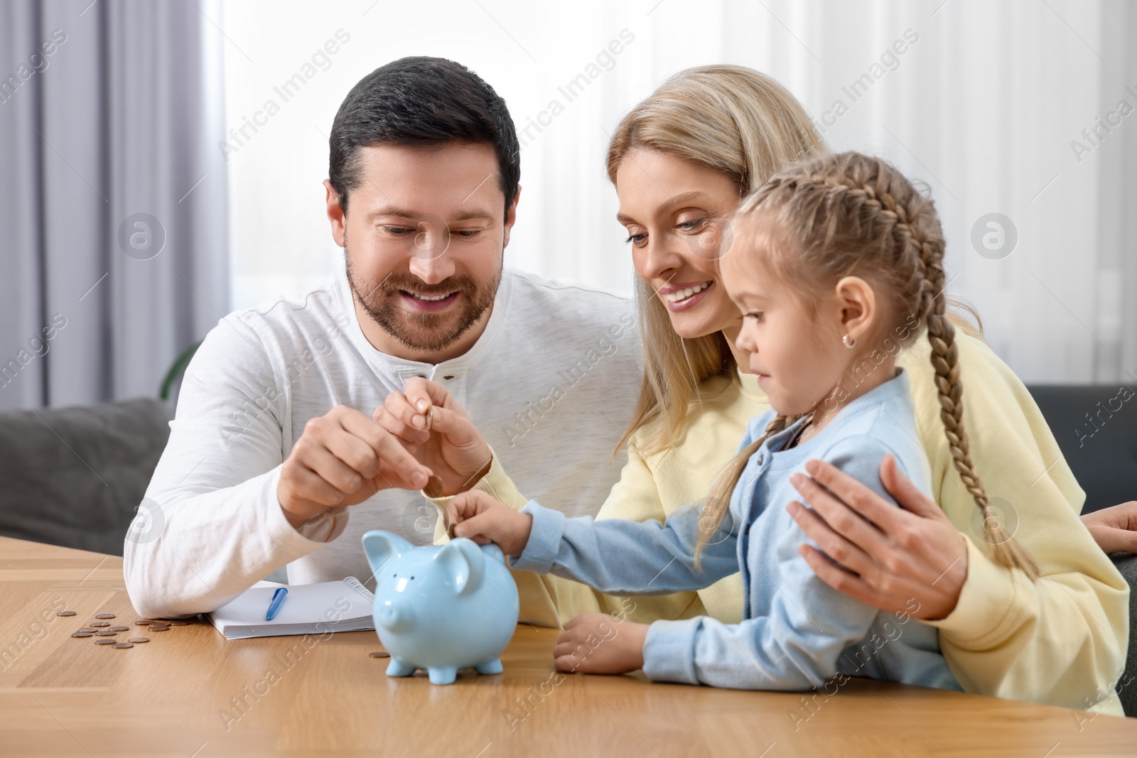 Photo of Planning budget together. Little girl with her family putting coins into piggybank at table indoors