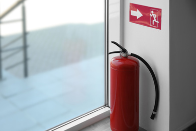Photo of Modern fire extinguisher and emergency exit sign near window indoors. Space for text