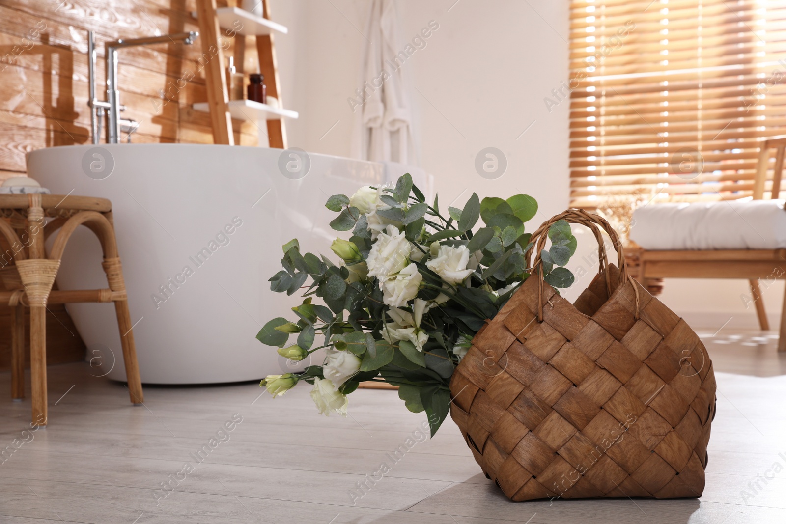 Photo of Stylish wicker basket with fresh eucalyptus branches and flowers on floor in bathroom